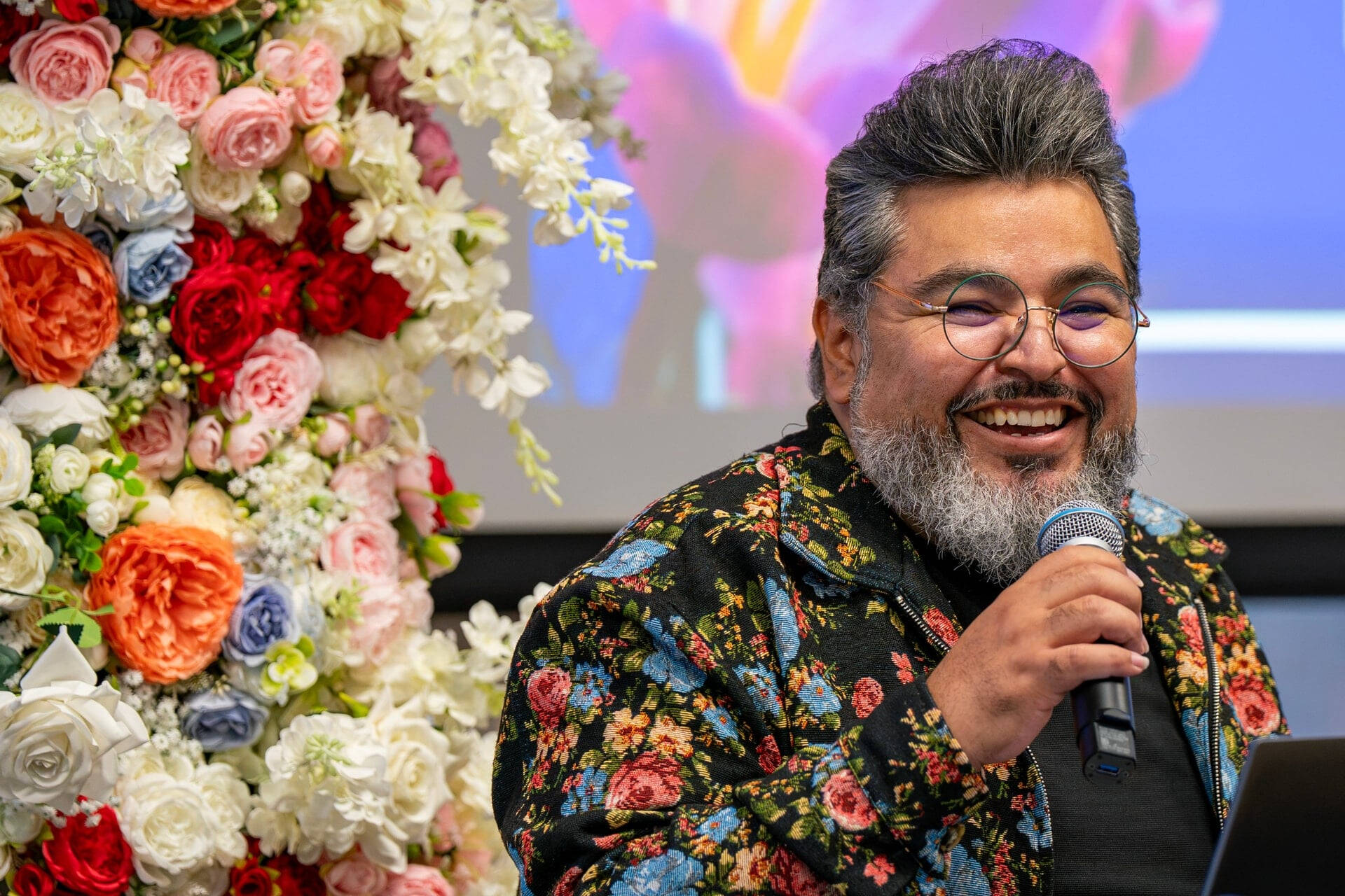 Tiburcio Lince holds a microphone, he is dressed in a floral jacket standing in front of a large flower display
